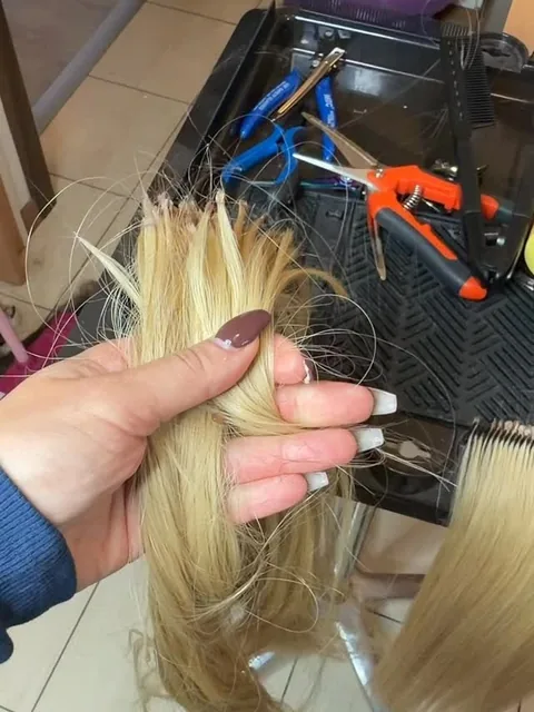Dłoń trzymająca przygotowane pasma blond przed aplikacją, w tle narzędzia na wózku fryzjerskim
